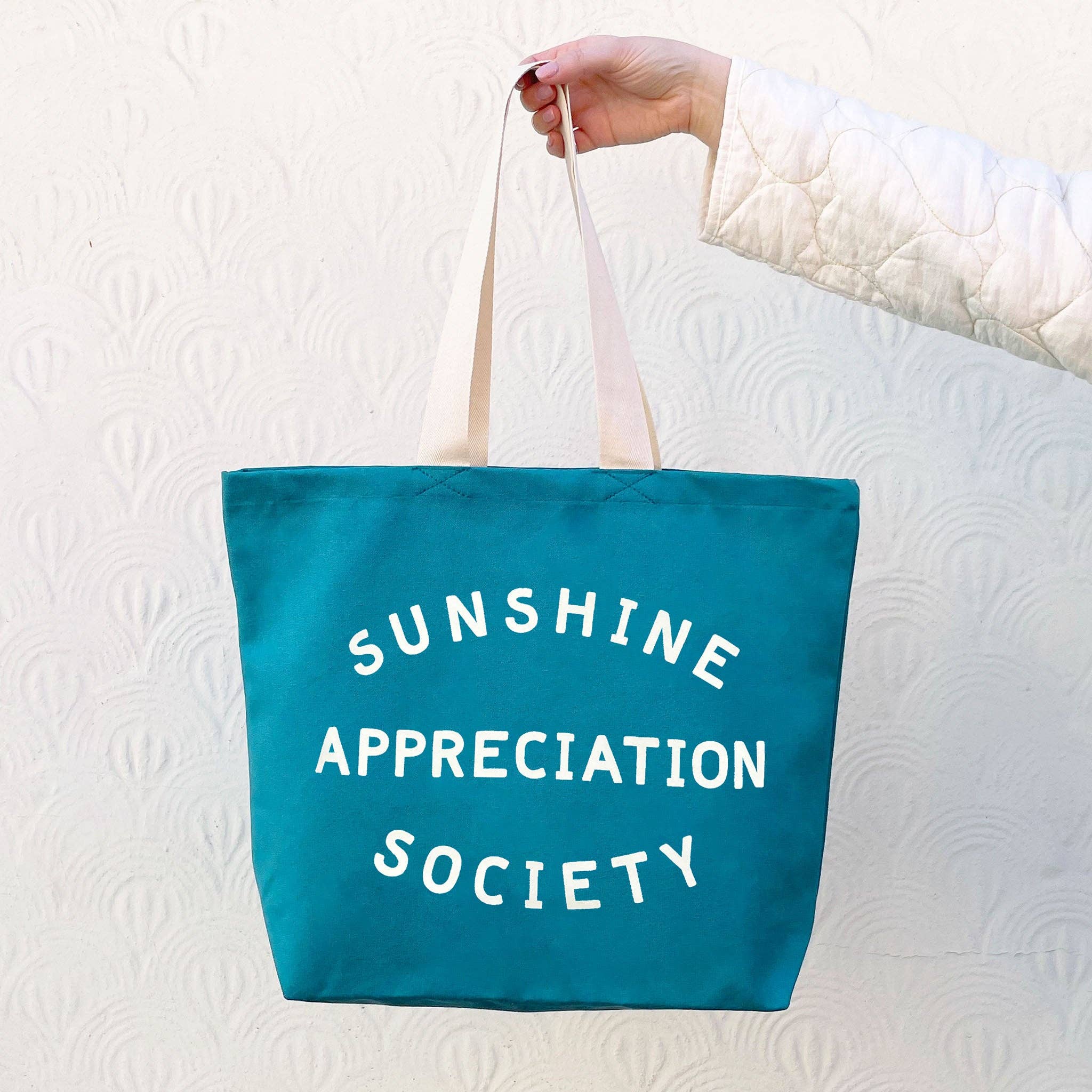 Teal tote bag with “Sunshine Appreciation Society” in white text, held by a person in a green dress at the beach
