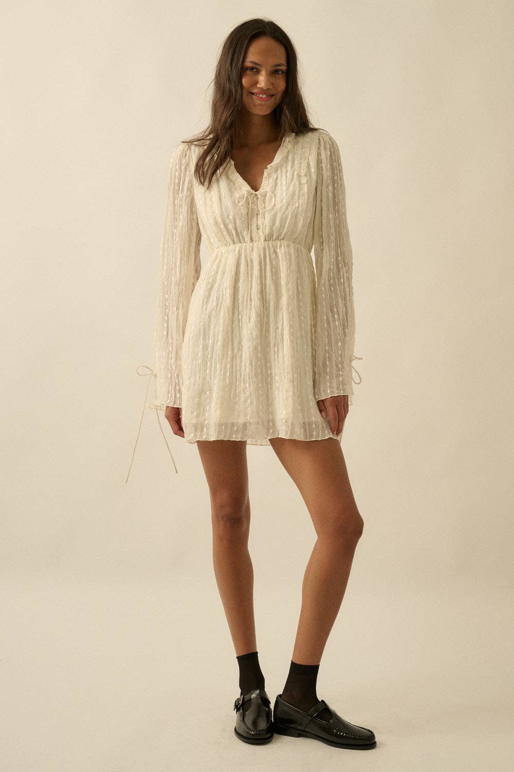 Woman wearing a cream mini dress with sheer long sleeves, button front, and tie details.