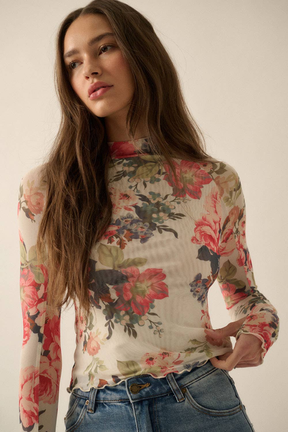 Model wearing a fitted sheer mesh long sleeve top with red, pink, and green floral print, paired with blue jeans.