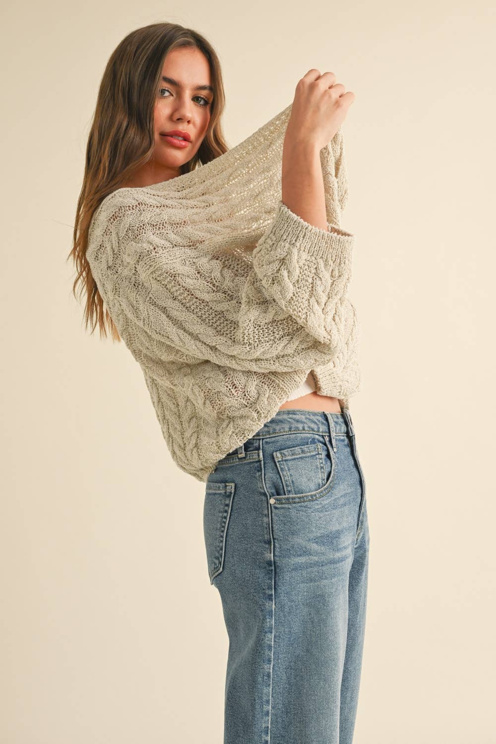 Woman wearing a loose cable knit cardigan with wide sleeves, paired with a white ribbed tank and blue denim jeans.