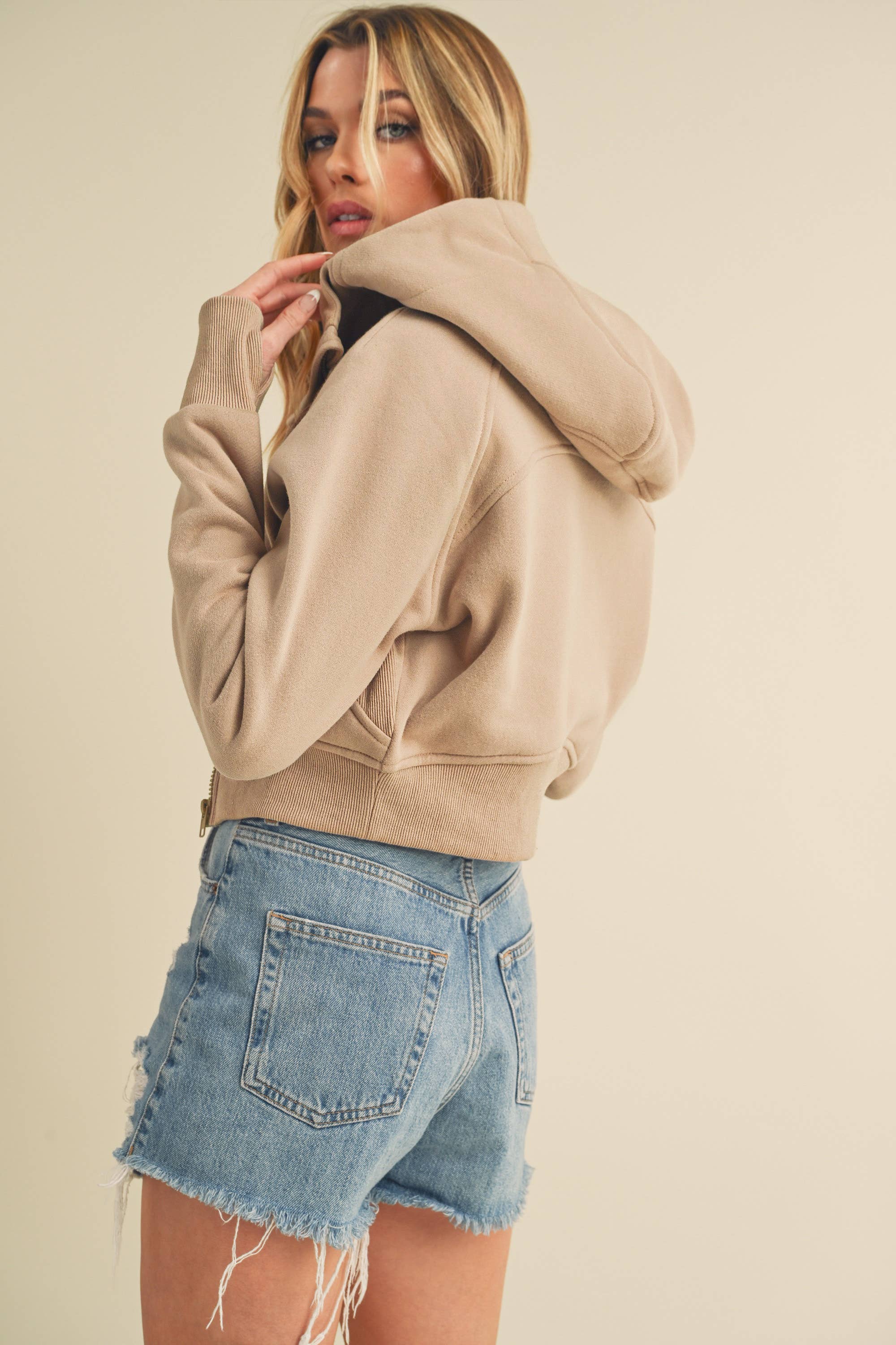 Model wearing a cropped zip-up hoodie with adjustable hood and ribbed cuffs, paired with distressed denim shorts.