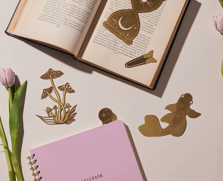A set of two brass metal page tabs in butterfly and mushroom shapes, packaged on a pastel pink and white wavy background.