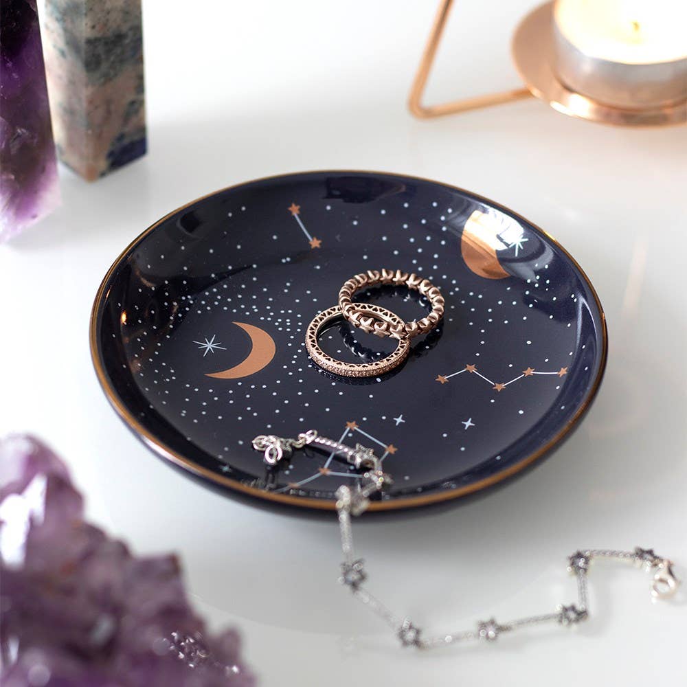 A round ceramic jewelry dish with a celestial design, including golden moons, stars, and constellations on a dark background.