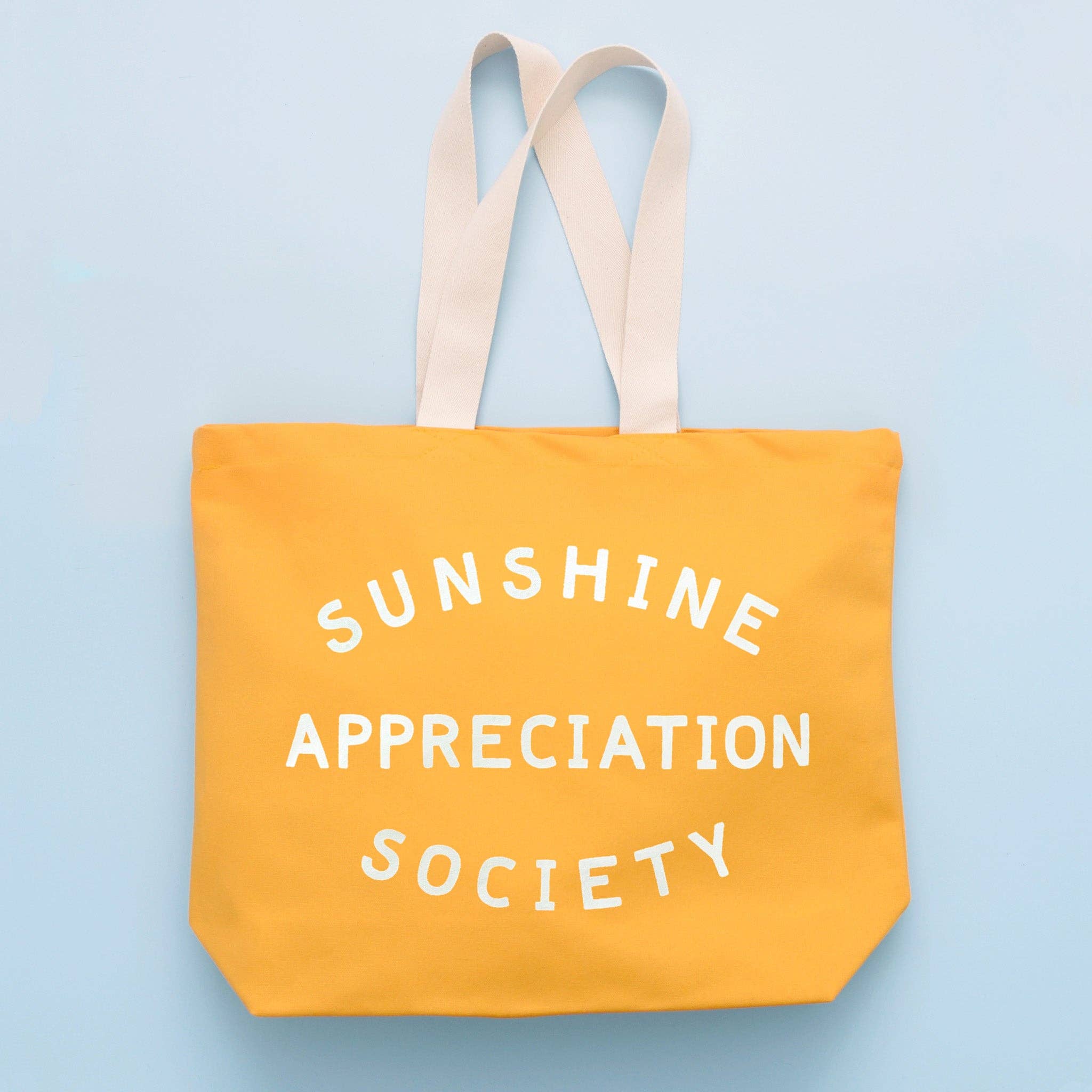 Yellow canvas tote bag with “Sunshine Appreciation Society” printed in white, held up by a hand in front of a sage green wall