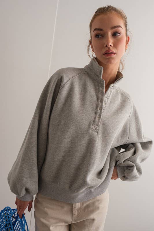 Woman wearing an oversized gray pullover sweatshirt with quarter snaps and voluminous sleeves, paired with beige pants.