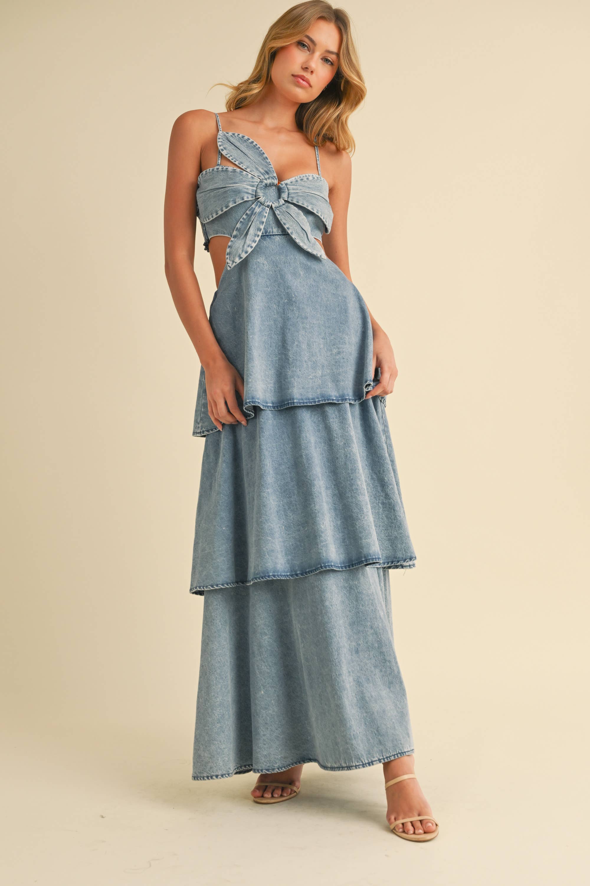 Woman wearing a denim maxi dress with a large front bow, side cutouts, spaghetti straps, and tiered skirt.