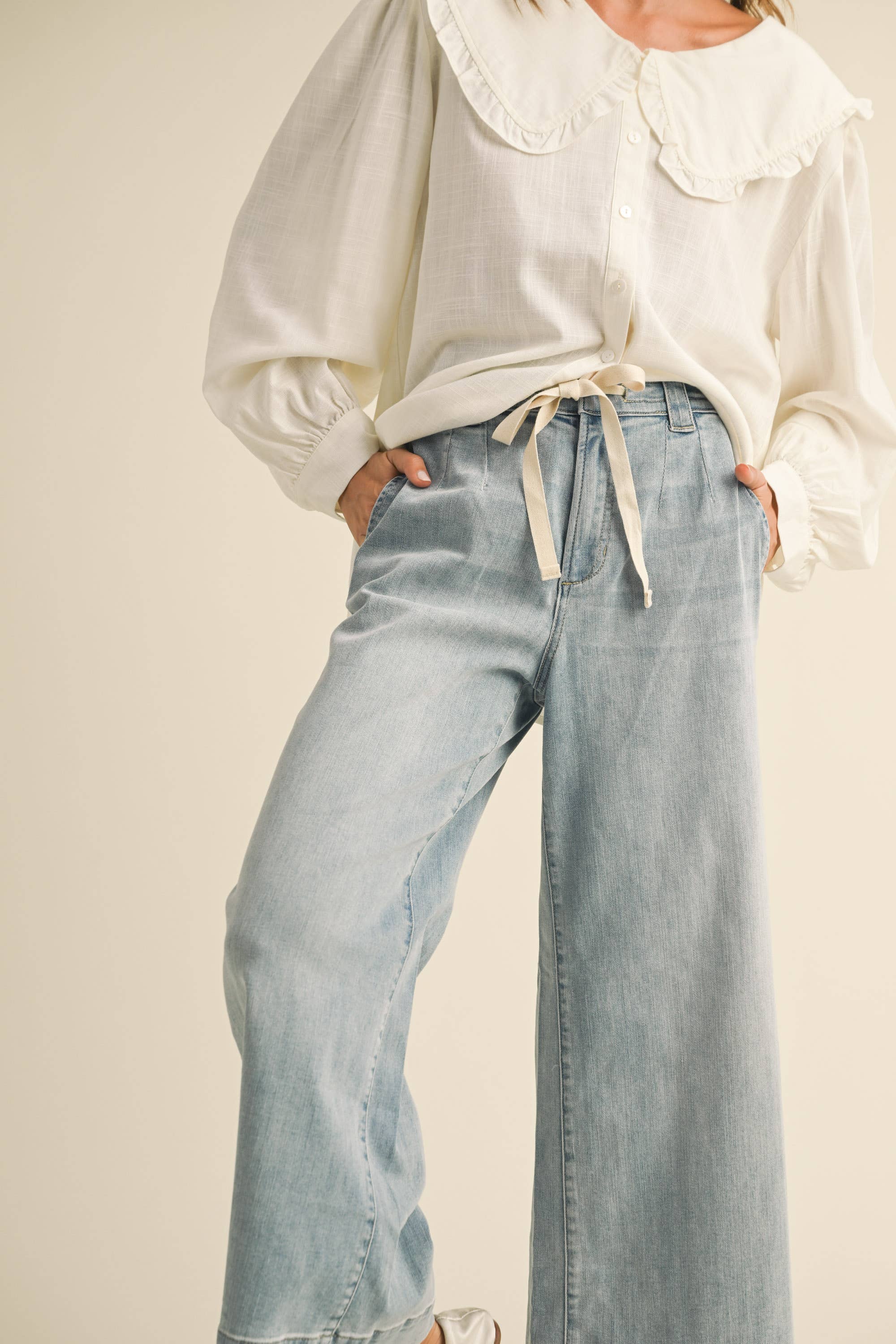 Woman wearing wide-leg light denim pants with a tie-front waist, paired with a white blouse and ballet flats.