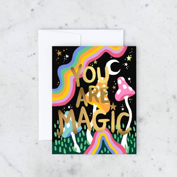 A greeting card featuring colorful rainbows, whimsical mushrooms, and celestial elements with the words “You Are Magic” in bold gold lettering.