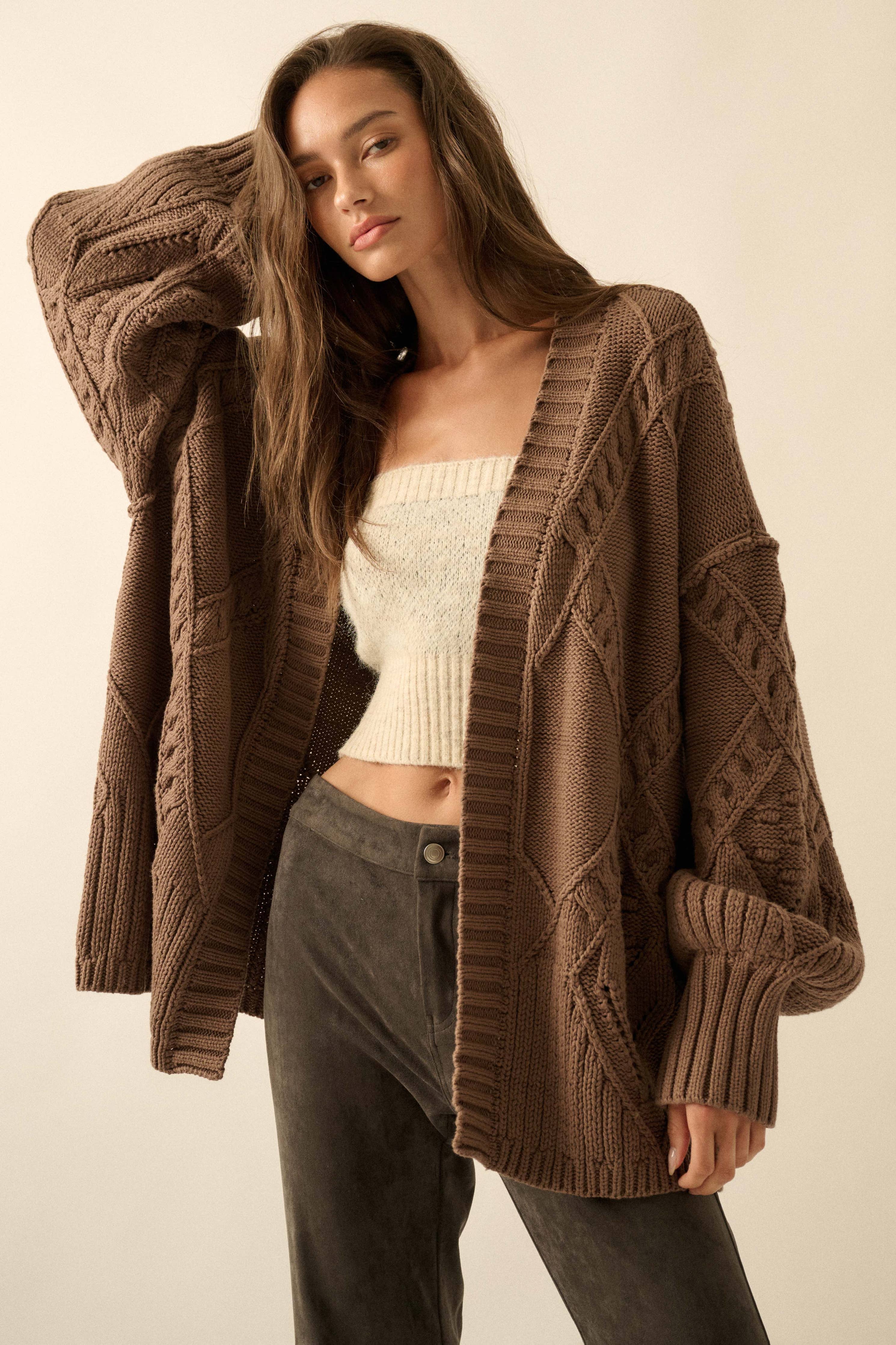 Woman wearing a brown cable knit cardigan over a white top and gray pants on a beige background