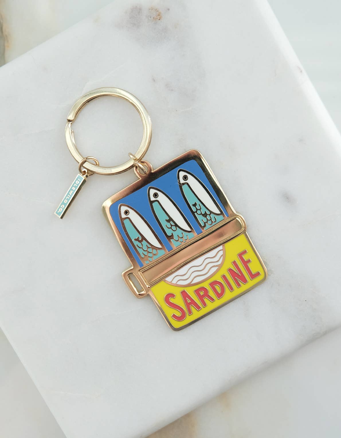 A brass and enamel keychain featuring a colorful design with sardines in a tin can, labeled 'SARDINE' on a yellow background, with a gold-colored ring.