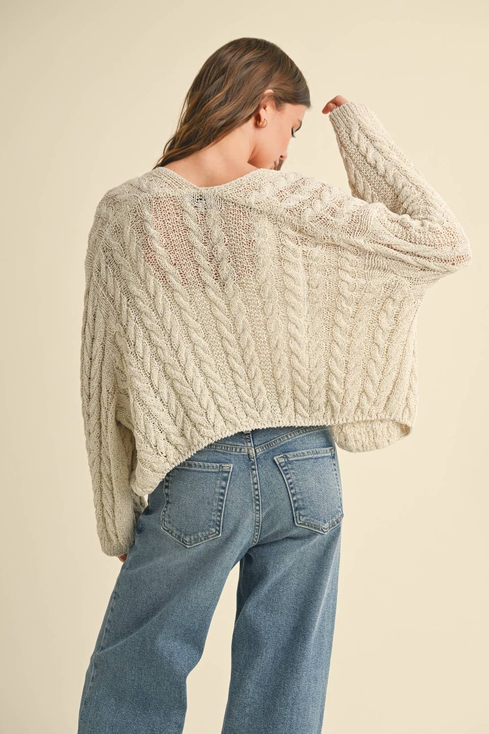 Woman wearing a loose cable knit cardigan with wide sleeves, paired with a white ribbed tank and blue denim jeans.