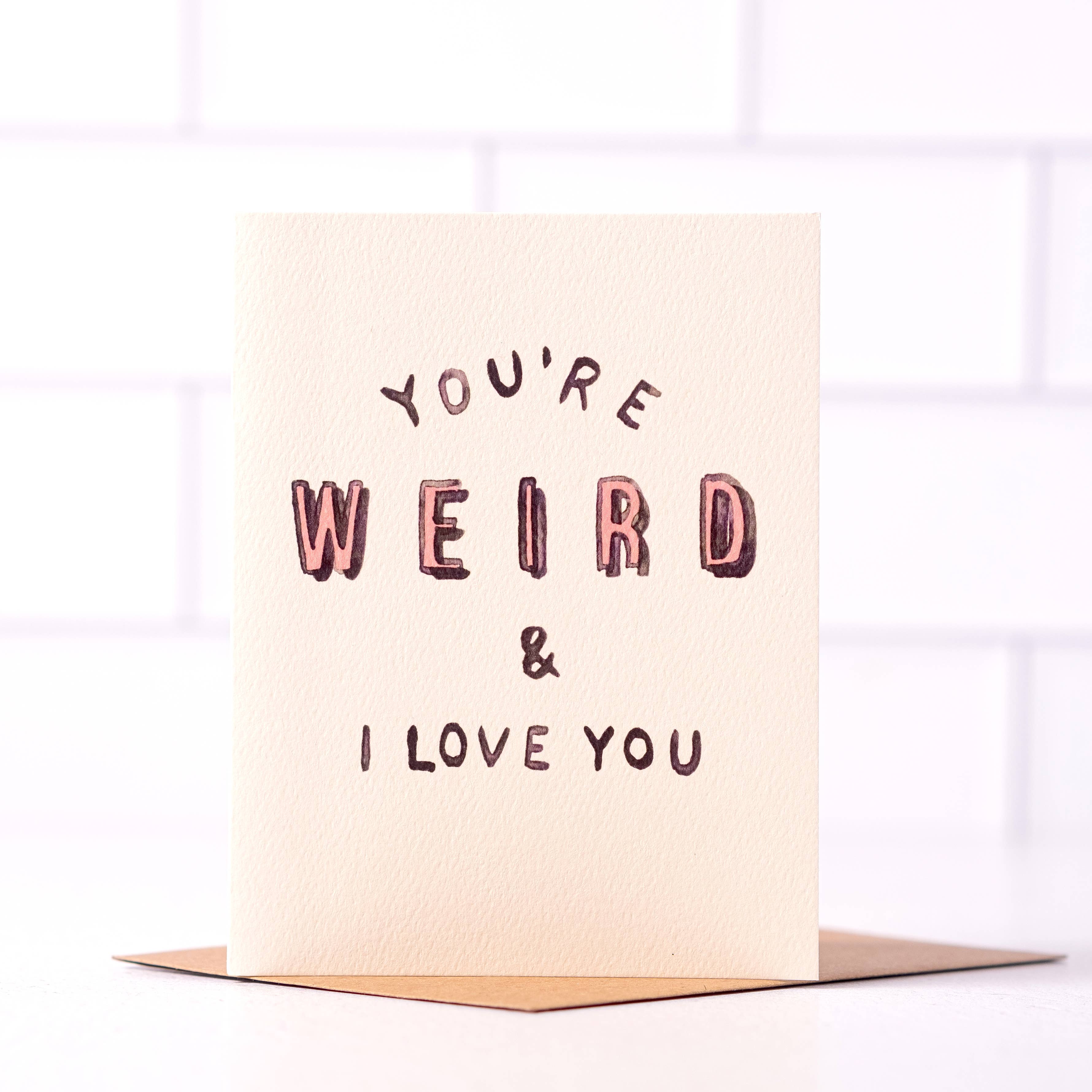 Off-white card with black and pink text saying ‘You’re Weird & I Love You,’ presented with a brown envelope.