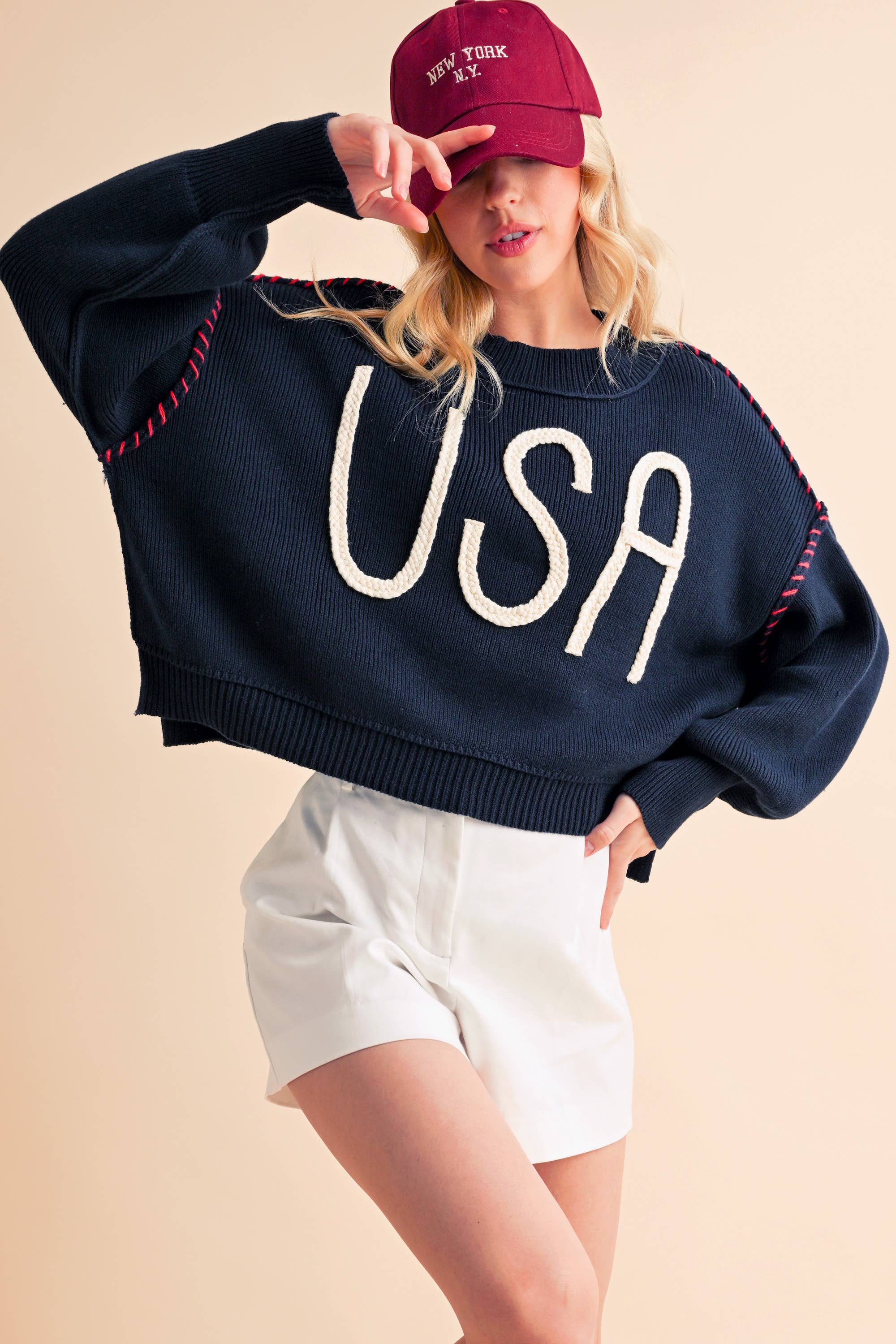 A woman wearing a navy sweater with the text 'USA' in white letters, paired with white shorts and a red cap.