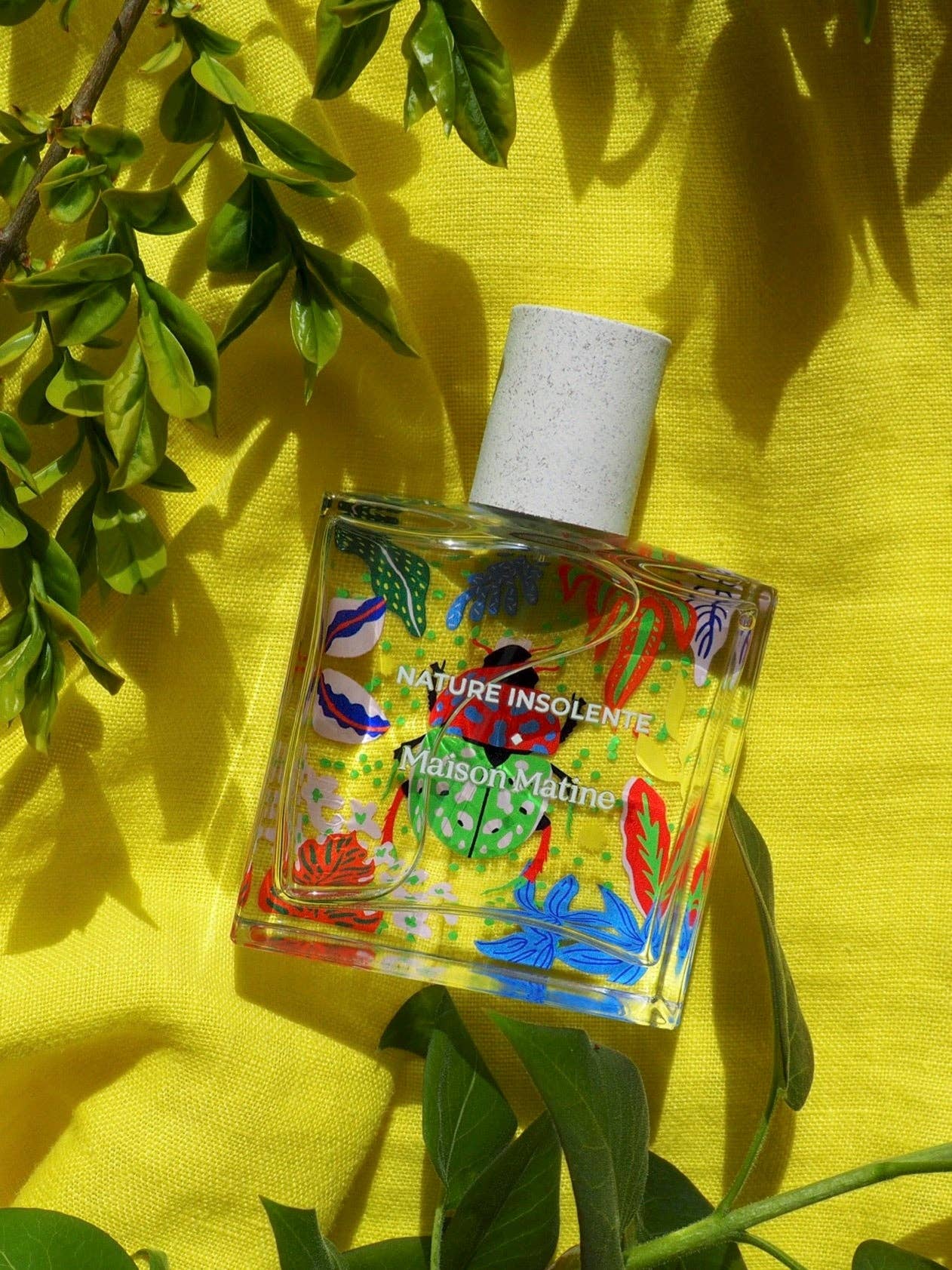 Maison Matine Nature Insolente perfume bottle with colorful leaf and beetle illustrations, placed on a vibrant yellow fabric and surrounded by green foliage.