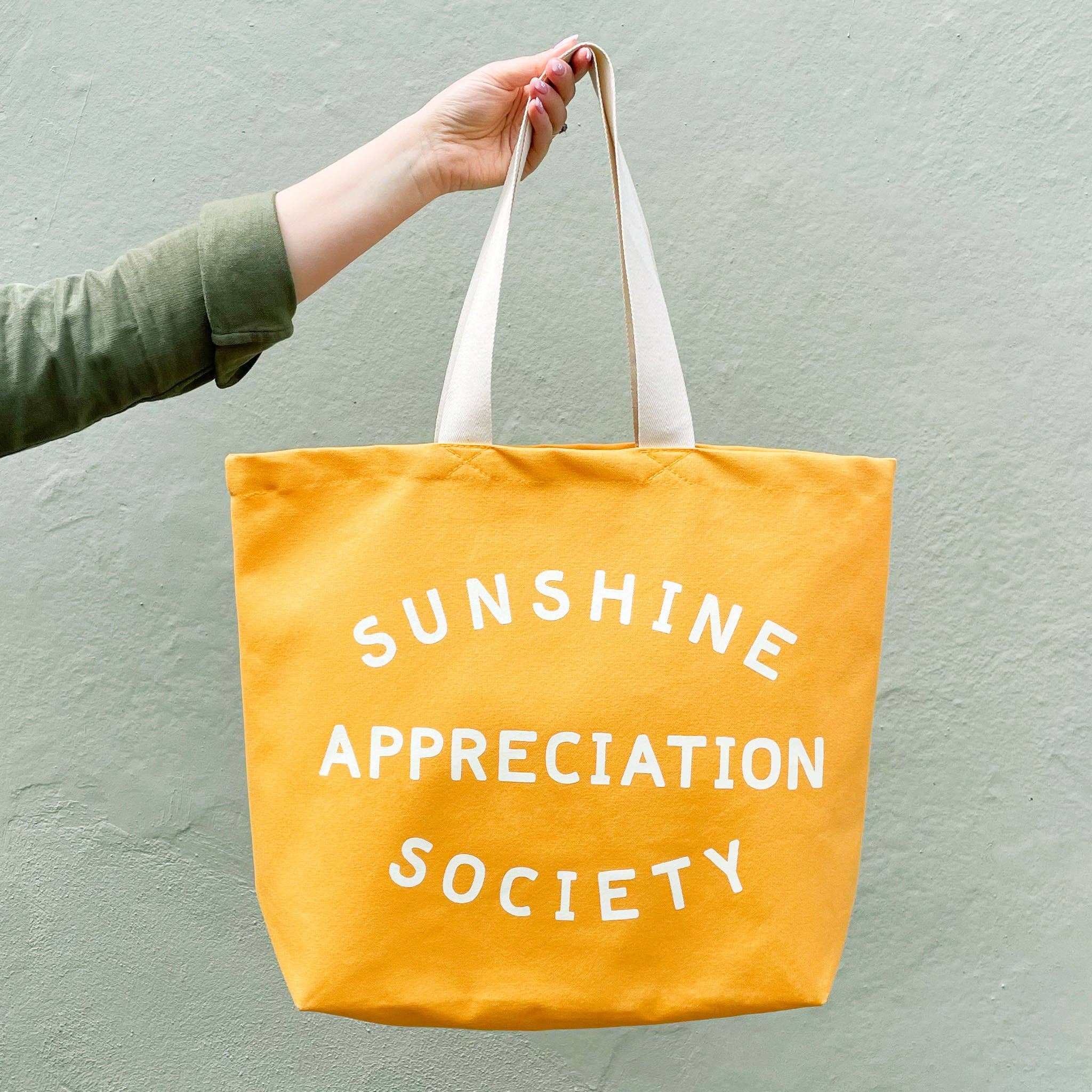 Yellow canvas tote bag with “Sunshine Appreciation Society” printed in white, held up by a hand in front of a sage green wall