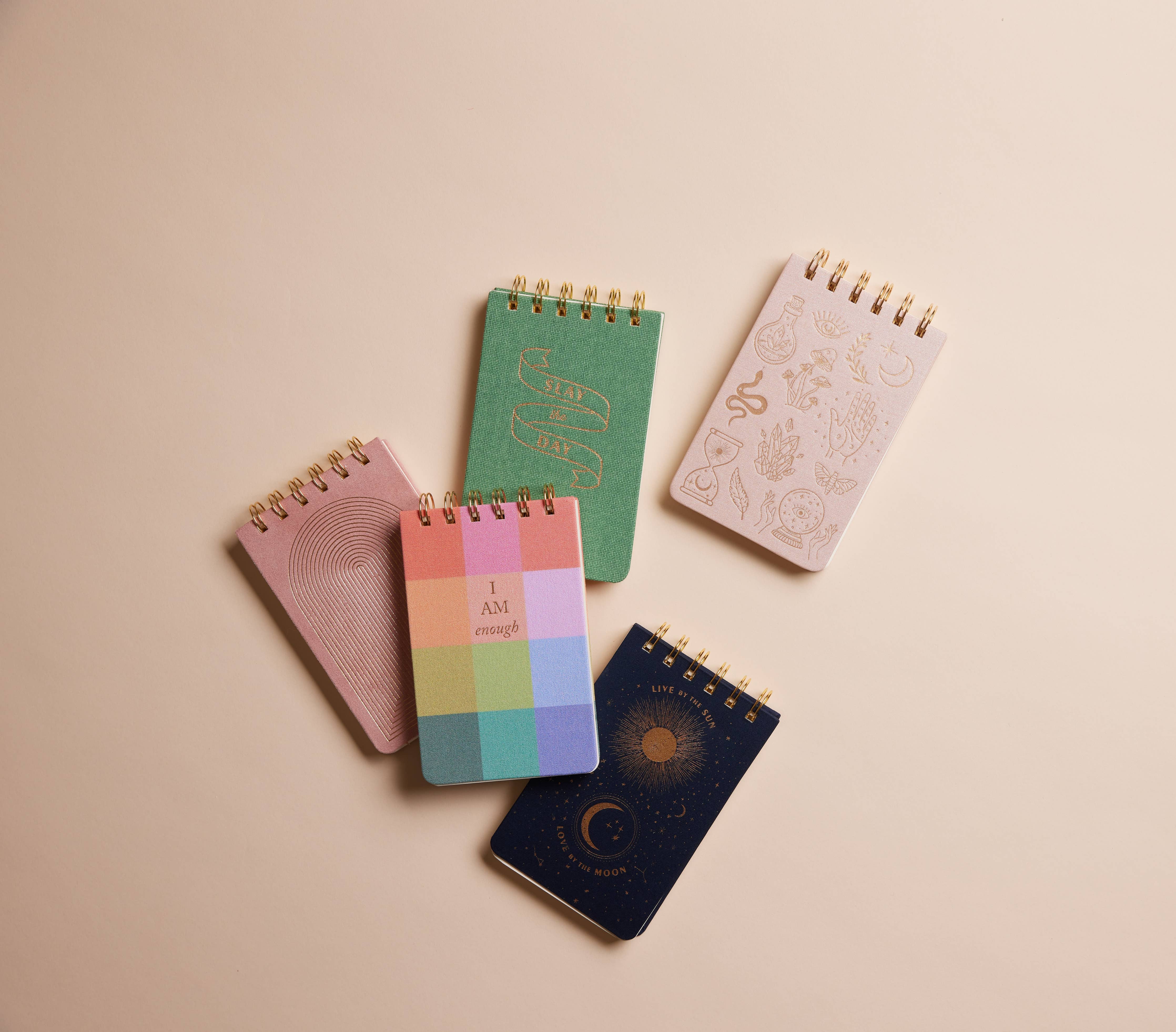 Five small spiral-bound notebooks with different covers on a beige background.