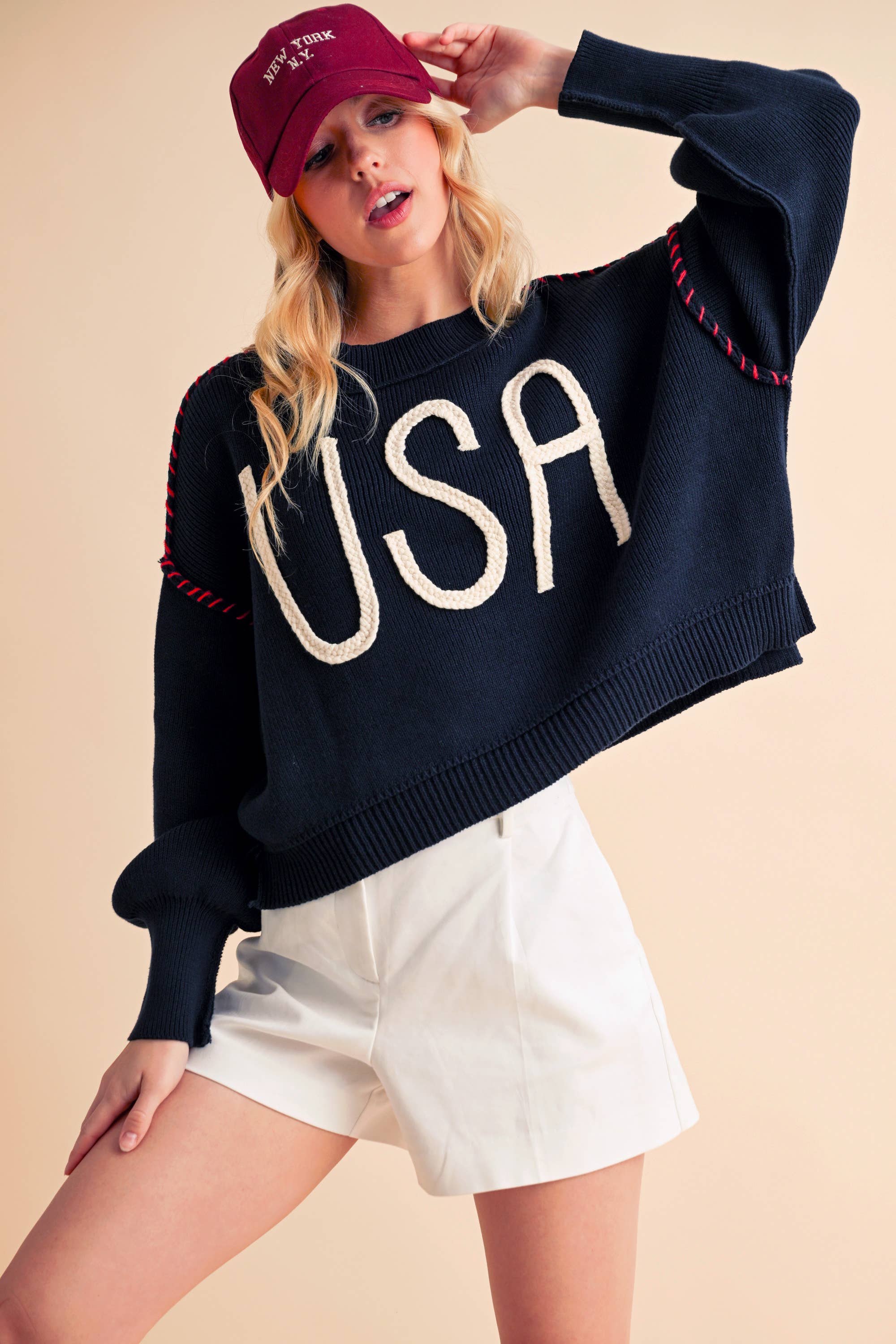 A woman wearing a navy sweater with the text 'USA' in white letters, paired with white shorts and a red cap.