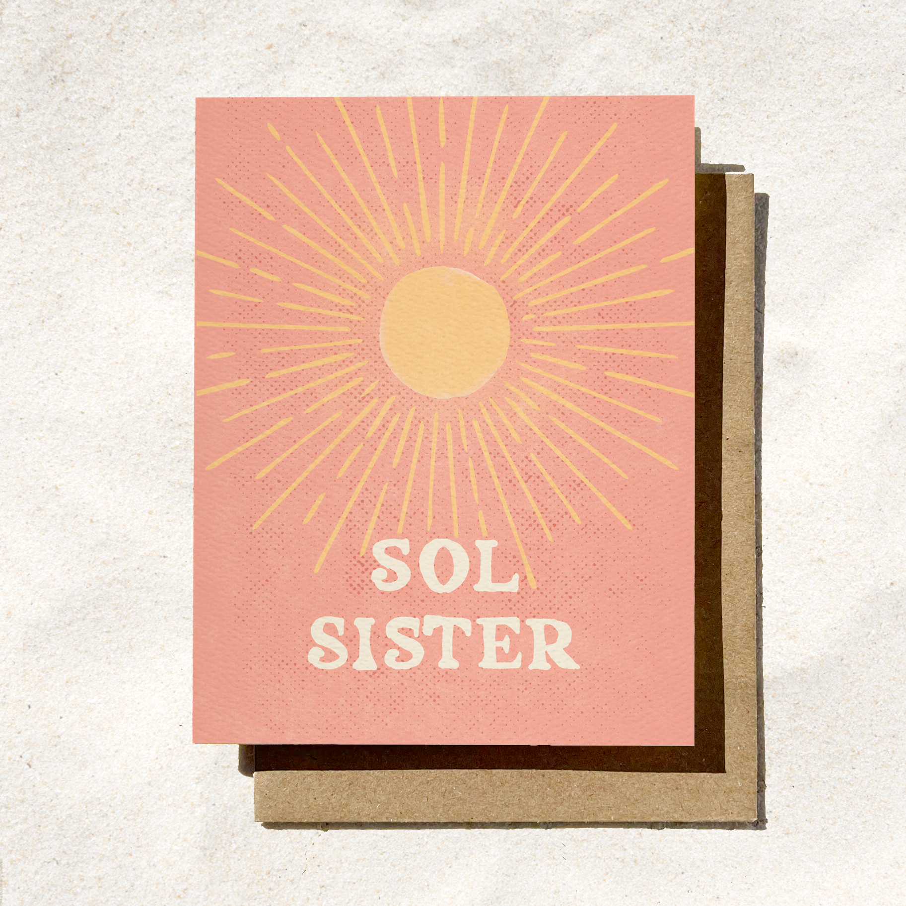 A peach-colored greeting card with a golden sunburst design and the phrase "Sol Sister," placed on a brown envelope on a sandy surface.