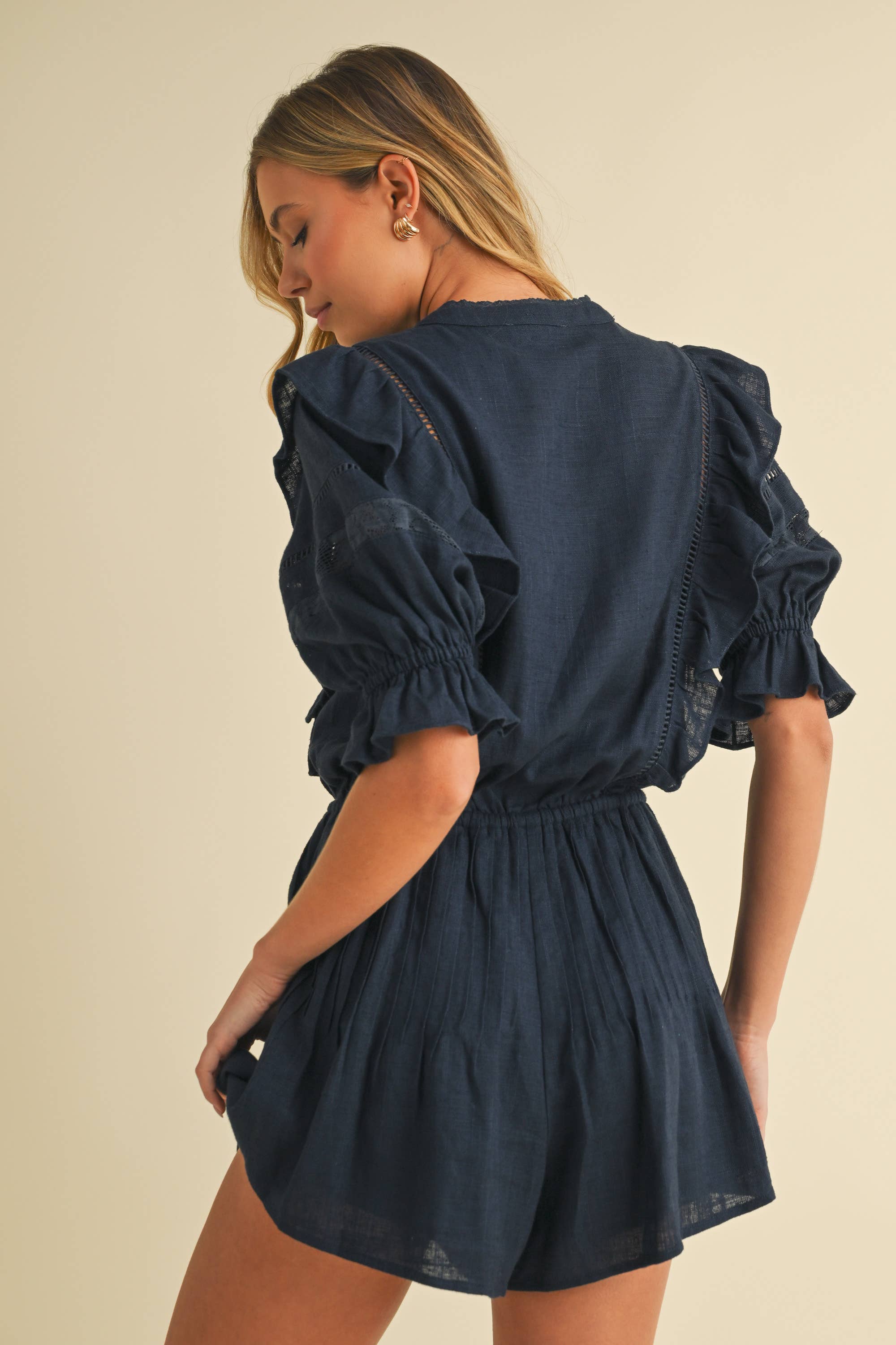 Woman wearing a navy cotton romper with puff sleeves, ruffle and lace details, and a drawstring waist.