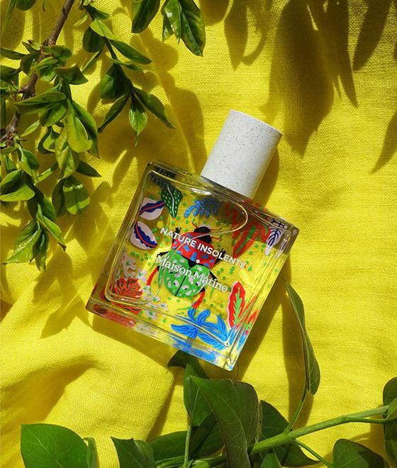 Maison Matine Nature Insolente perfume bottle with colorful leaf and beetle illustrations, placed on a vibrant yellow fabric and surrounded by green foliage.