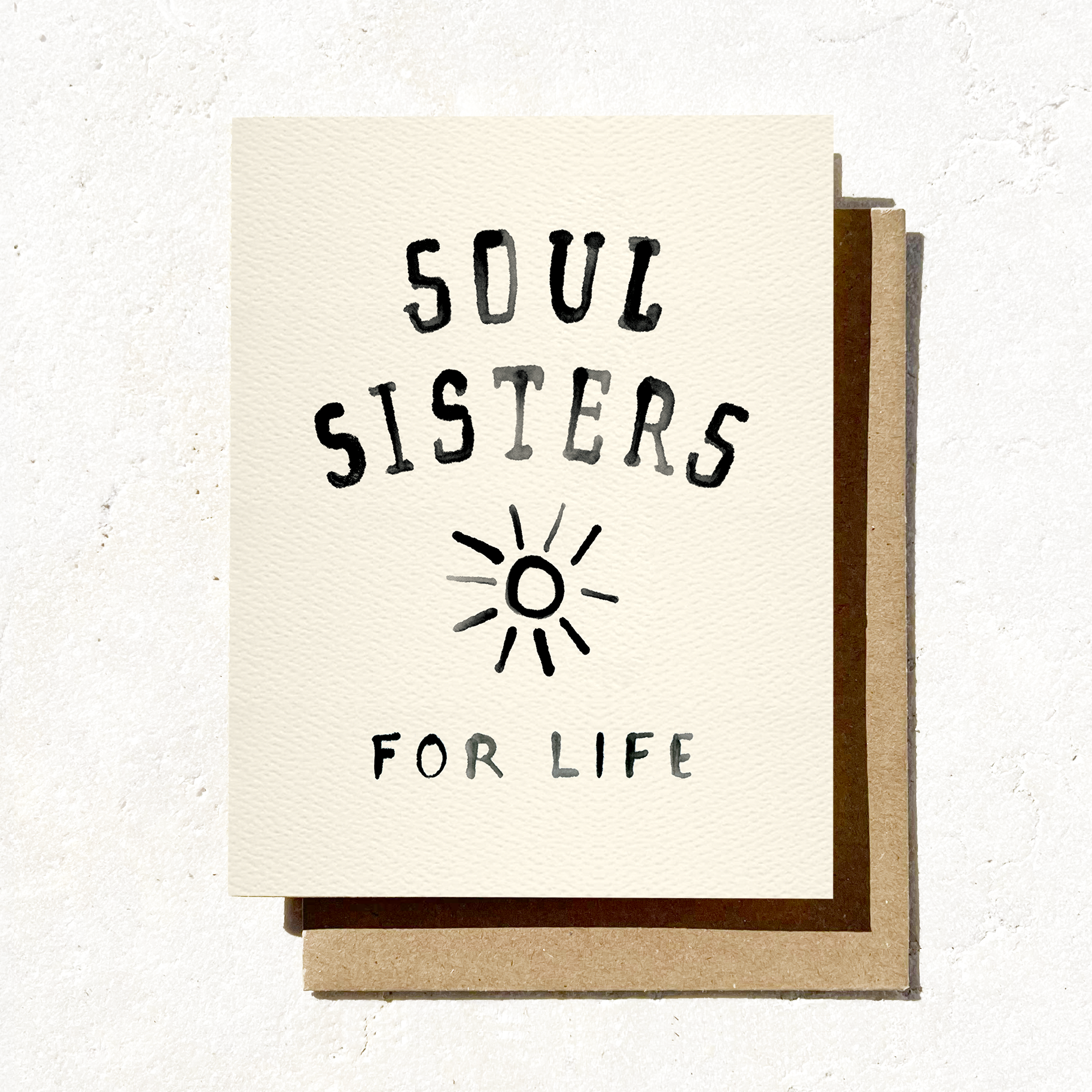 A cream-colored greeting card with "Soul Sisters for Life" in black hand-lettering and a sun doodle, placed on a brown envelope on a sandy surface.