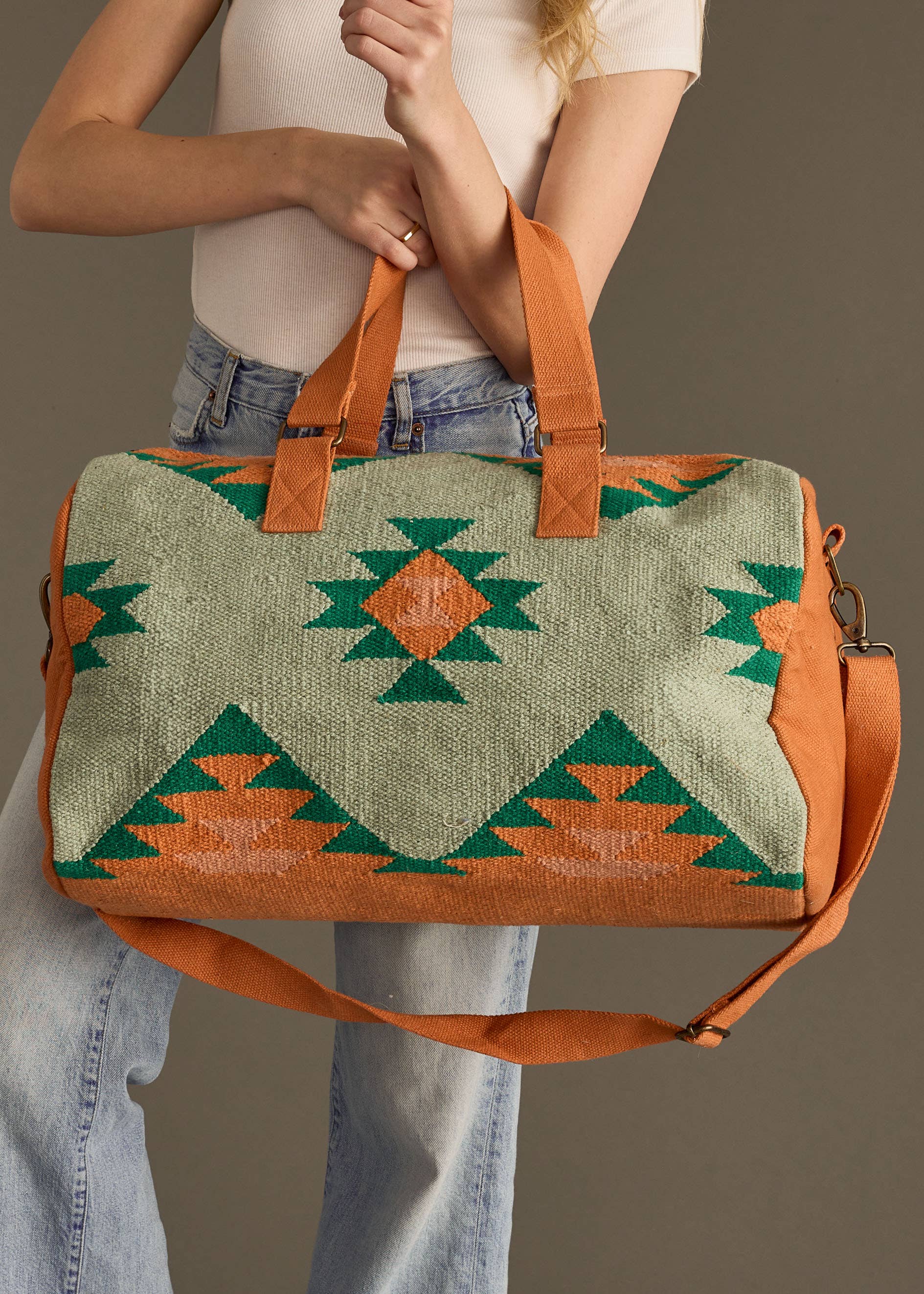 Sage green and rust weekender bag with geometric tribal pattern in green and orange, held by woman in jeans and white tank