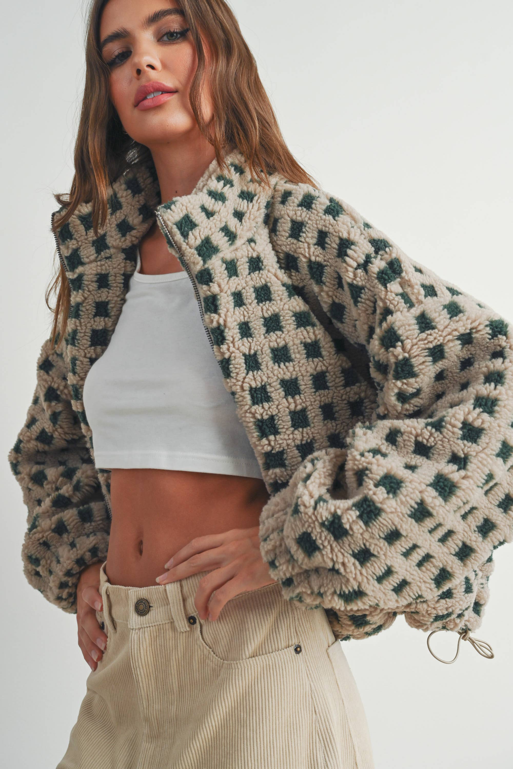 Woman wearing a beige and green checkered fleece jacket with a zip closure, paired with a white crop top and beige corduroy pants.
