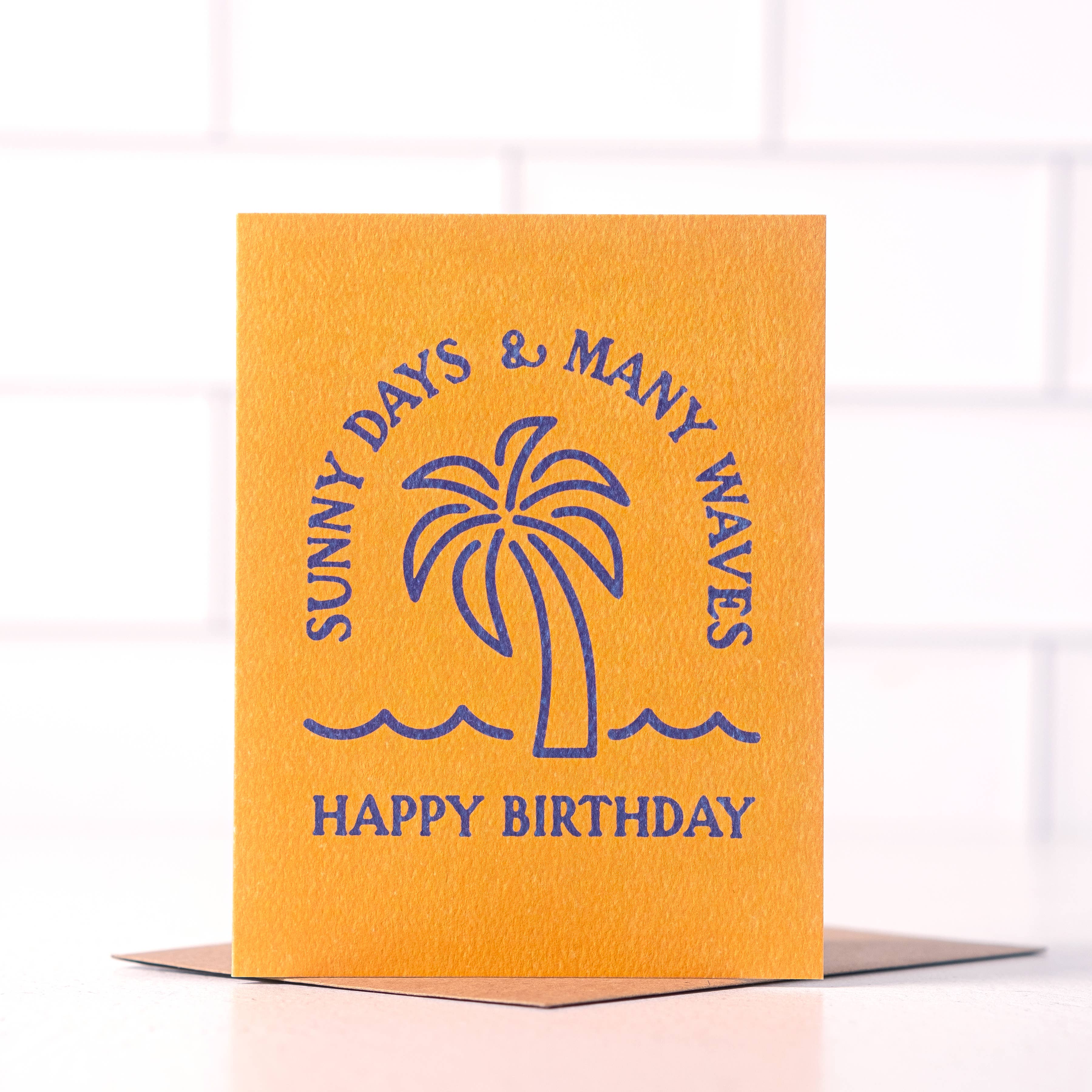 Orange birthday card with blue text saying ‘Sunny Days & Many Waves, Happy Birthday,’ featuring a palm tree illustration.