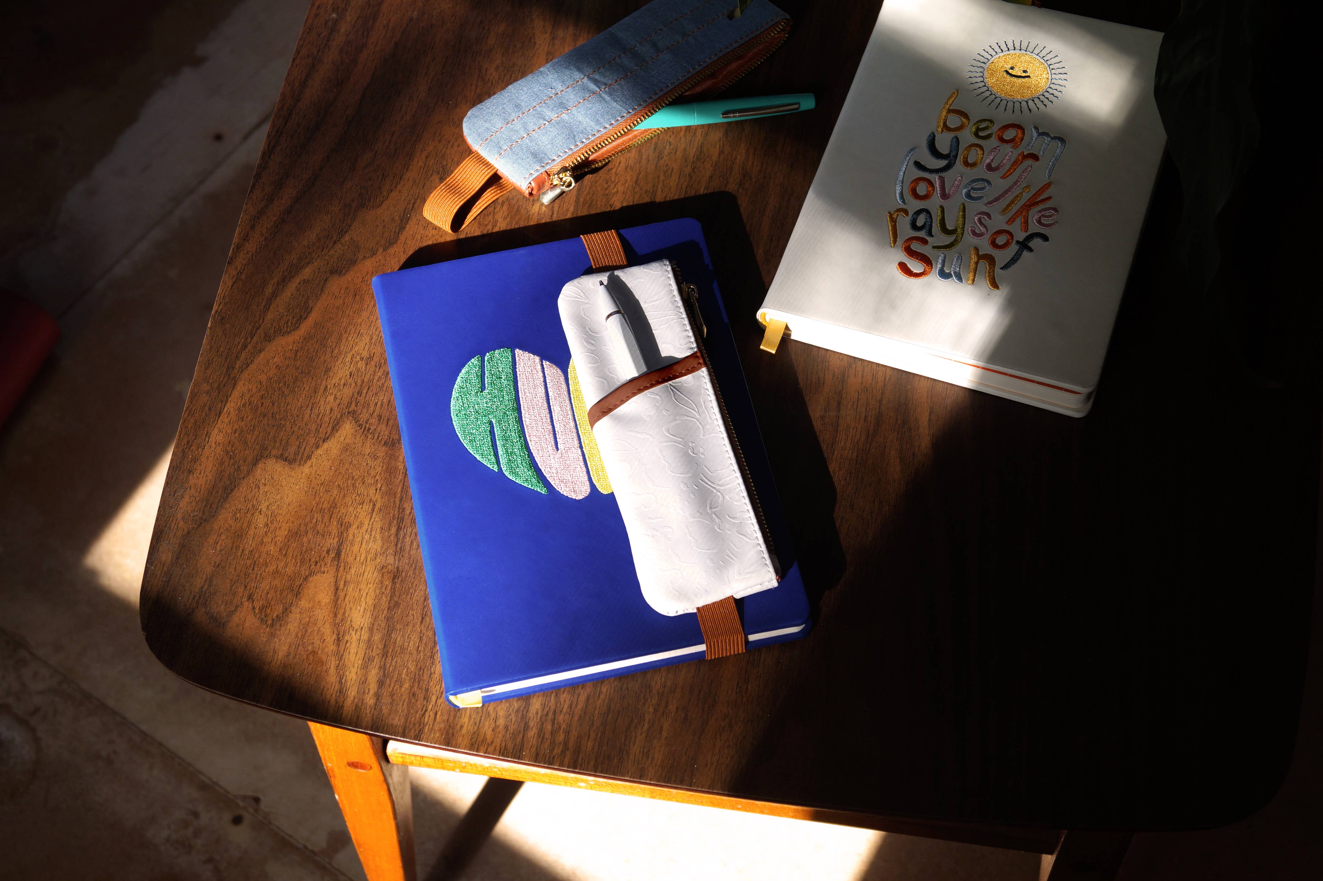 A white vegan leather notebook pouch with an embossed floral design, tan accents, and a gold zipper, attached to its packaging.