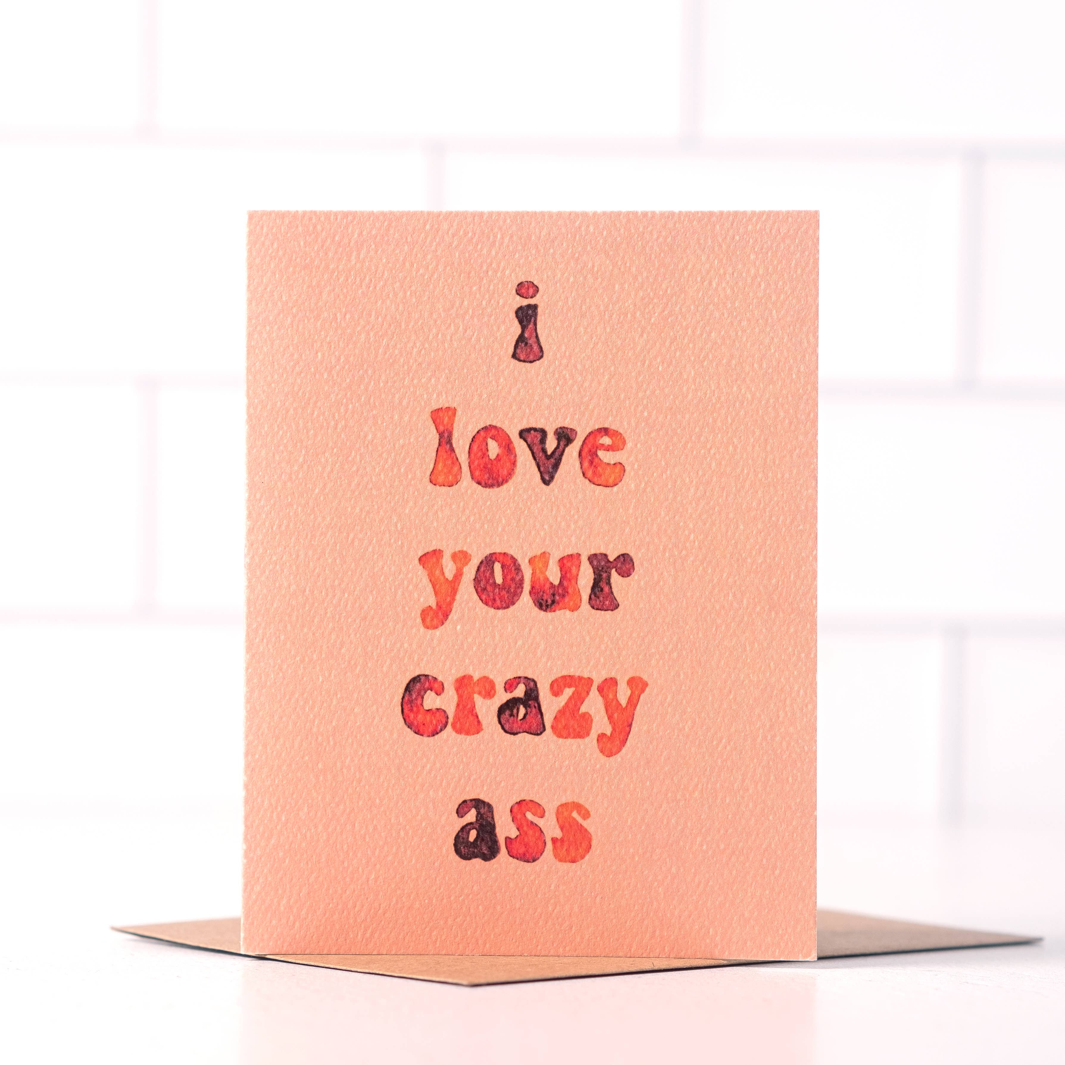 A blush-colored greeting card with "I love your crazy ass" in red and orange watercolor-style text.