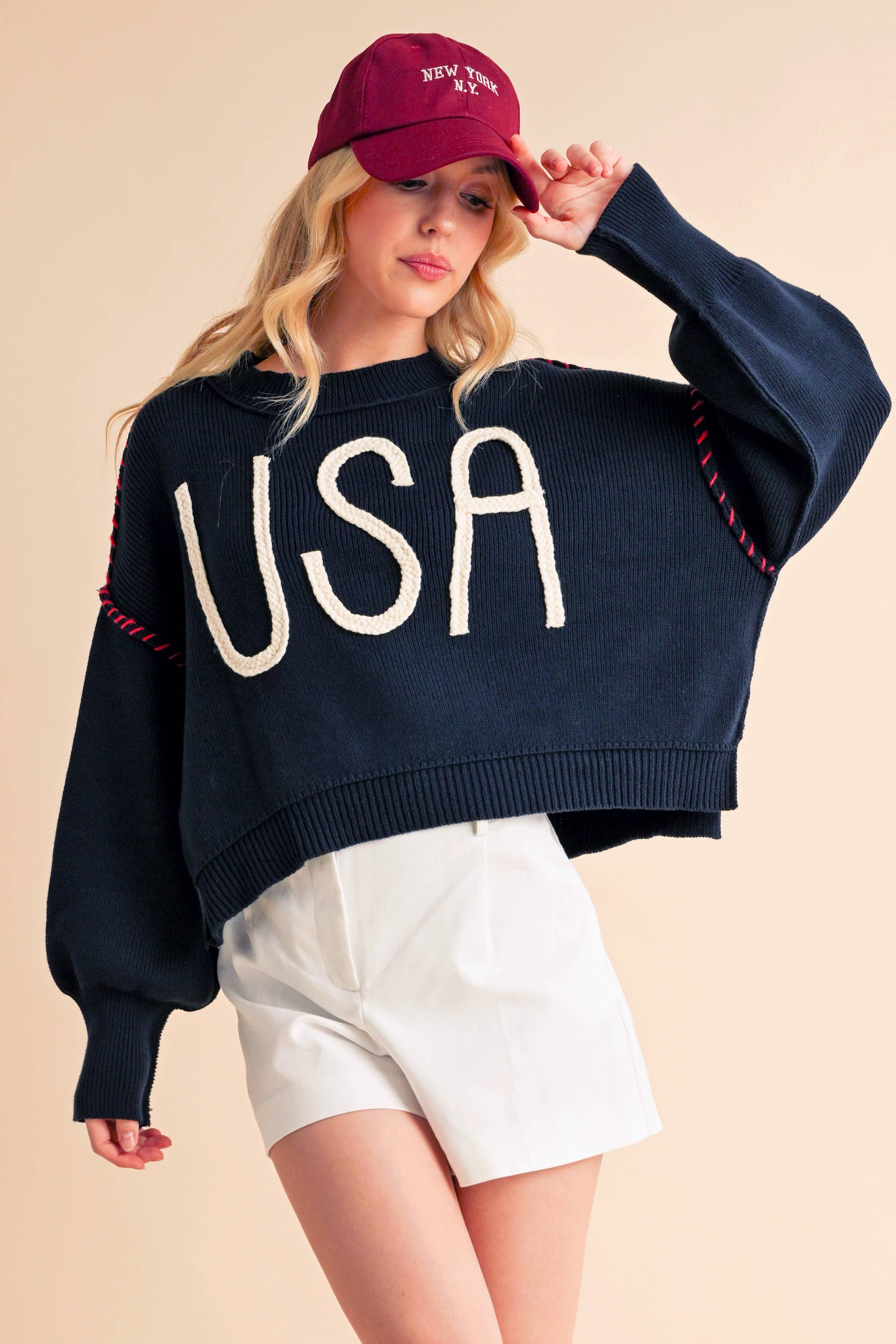 A woman wearing a navy sweater with the text 'USA' in white letters, paired with white shorts and a red cap.