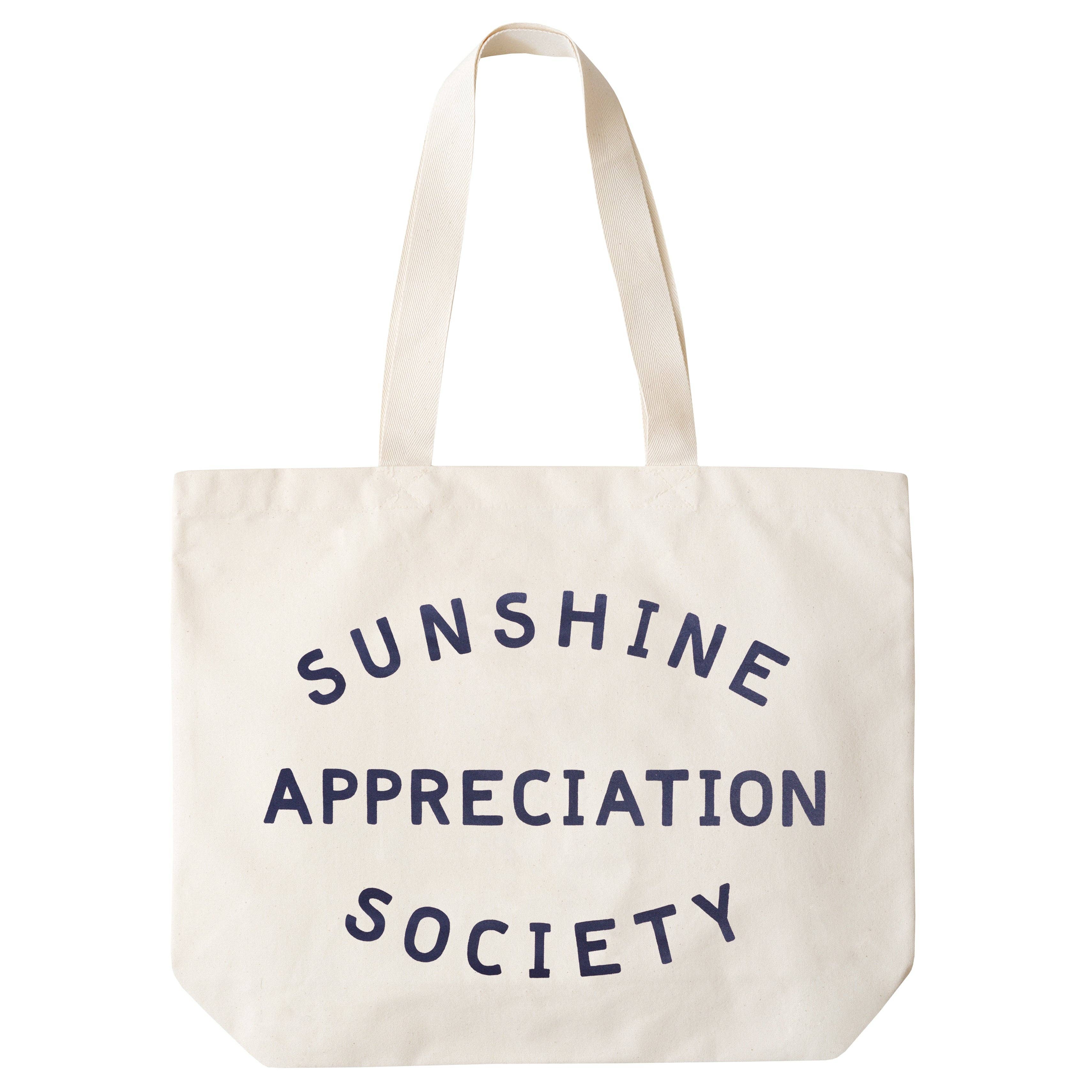 Cream canvas tote bag with “Sunshine Appreciation Society” printed in navy, filled with flip-flops, sunglasses, and travel essentials on a pink background