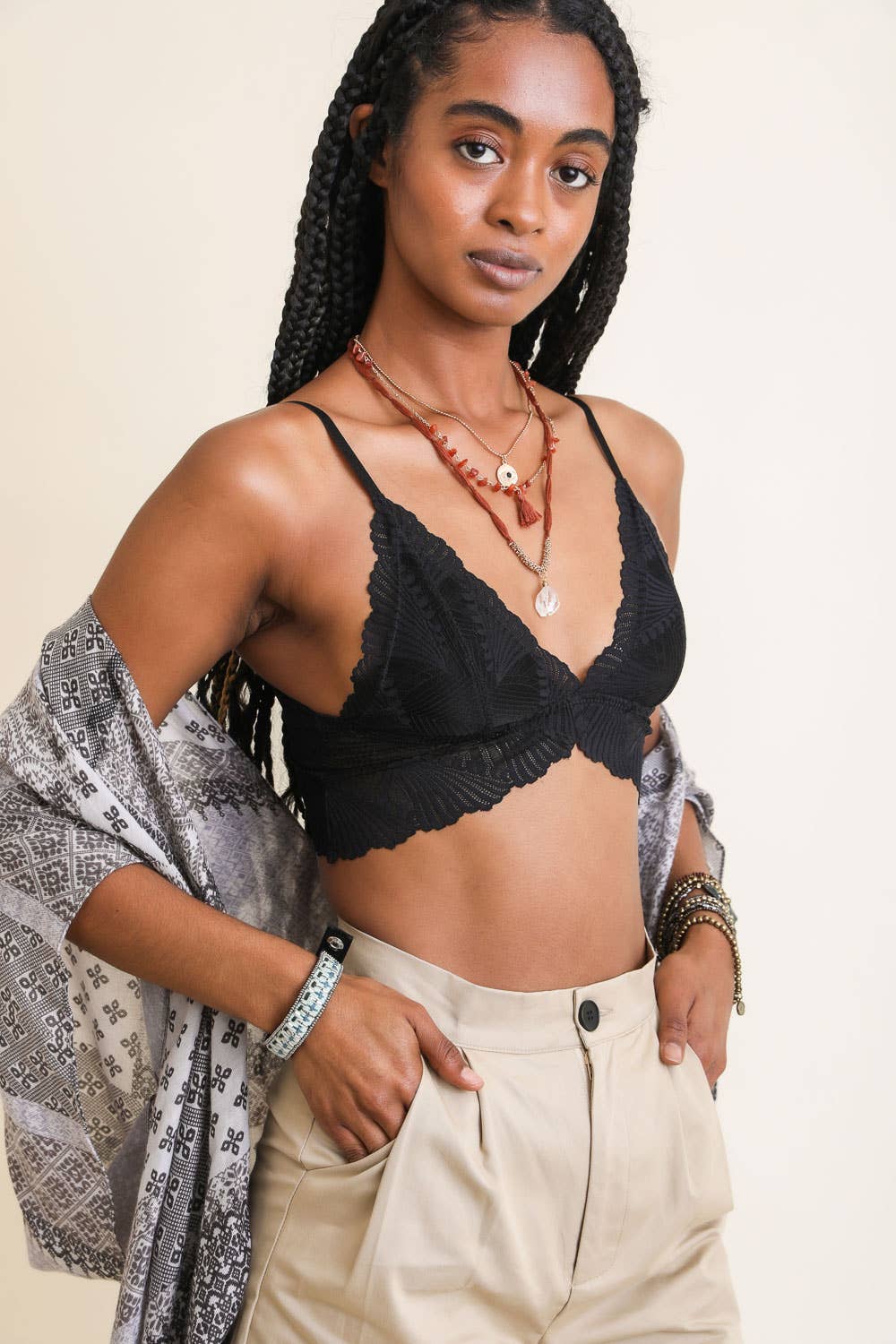 Model wearing a scalloped-edge lace bralette in burgundy and blue tones, styled with layered necklaces and velvet pants.