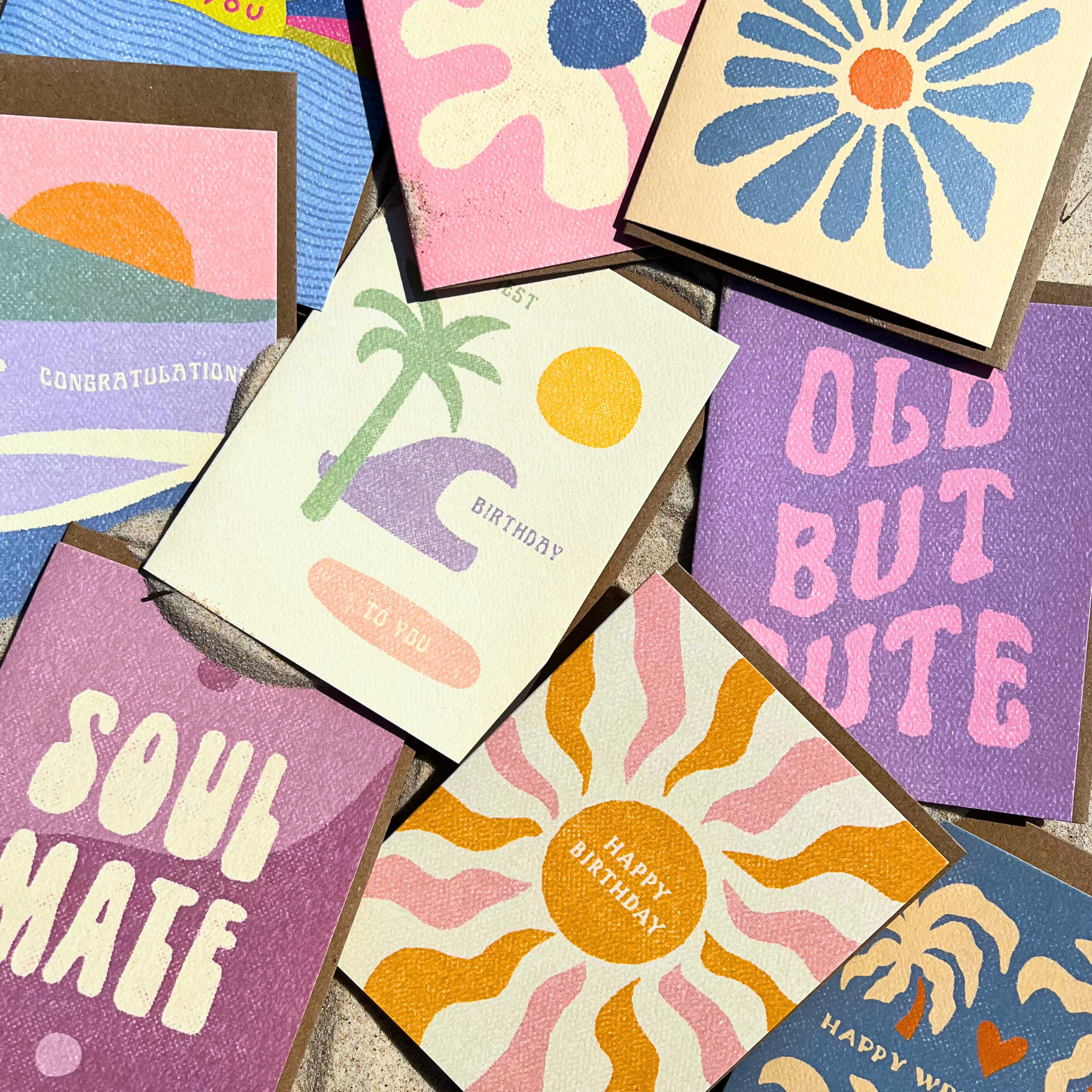 A cream-colored greeting card with "Soul Sisters for Life" in black hand-lettering and a sun doodle, placed on a brown envelope on a sandy surface.