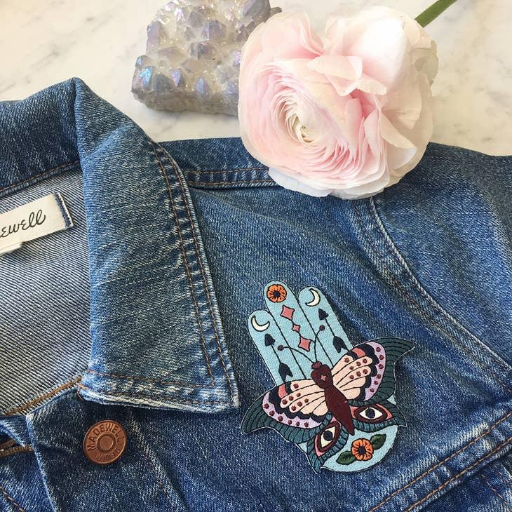 Large embroidered patch displaying a Hamsa integrated with butterfly, evil eyes, crescent moons, arrows, and floral motifs in a vibrant boho color palette.