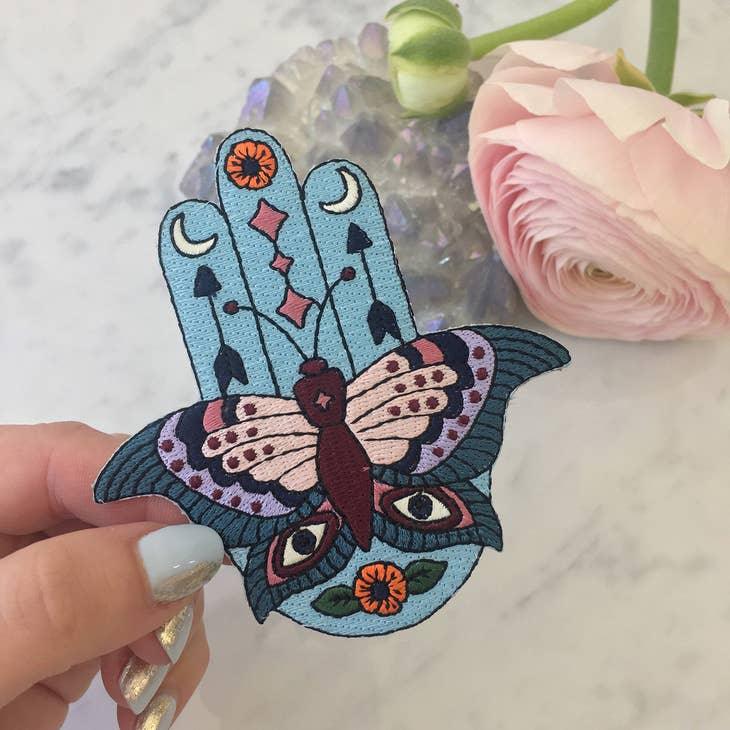 Large embroidered patch displaying a Hamsa integrated with butterfly, evil eyes, crescent moons, arrows, and floral motifs in a vibrant boho color palette.