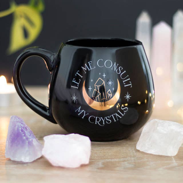 Black mug with ‘Let Me Consult My Crystals’ text and a gold foil crescent moon and crystal design, placed among crystals.