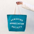 Teal tote bag with “Sunshine Appreciation Society” in white text, held by a person in a green dress at the beach