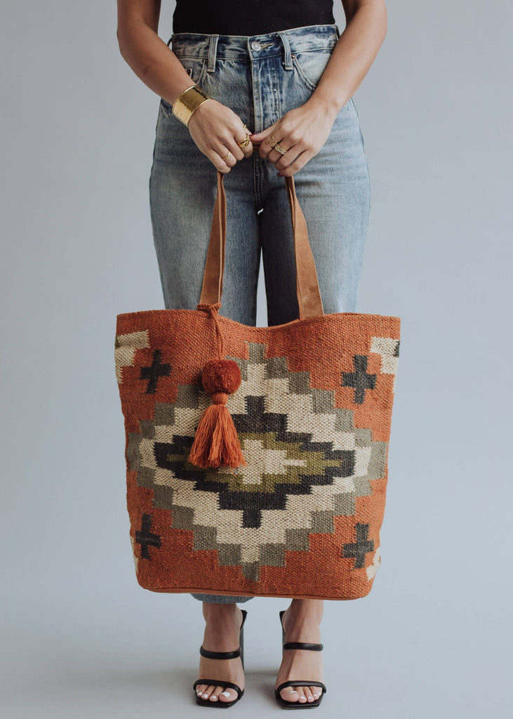 Woman holding a rust and olive green diamond-patterned woven tote with tassel
