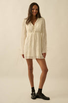 Woman wearing a cream mini dress with sheer long sleeves, button front, and tie details.