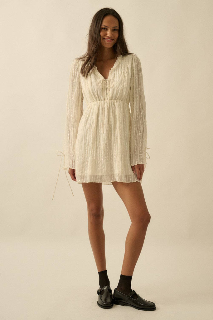 Woman wearing a cream mini dress with sheer long sleeves, button front, and tie details.