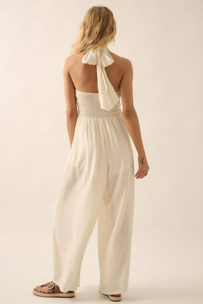 Woman wearing a white halter jumpsuit with tie-back neckline and wide-leg pants, styled with sandals.