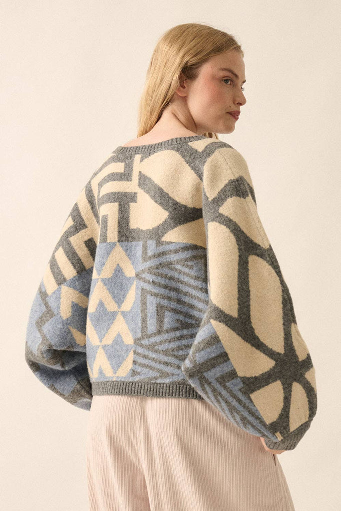 Model wearing a patterned knit sweater with bold geometric designs in neutral and contrasting tones, styled with casual bottoms.