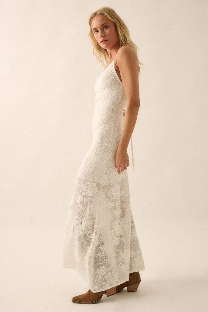 Woman wearing a sheer white lace maxi dress with spaghetti straps and floral detailing.