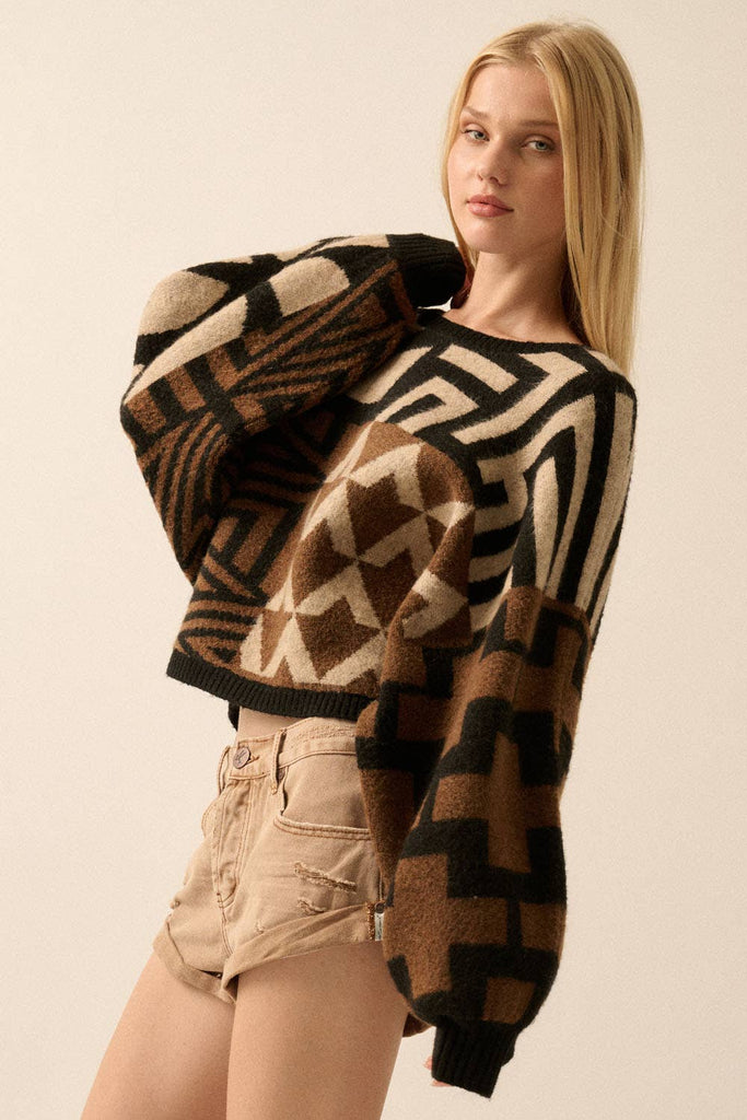 Model wearing a patterned knit sweater with bold geometric designs in neutral and contrasting tones, styled with casual bottoms.