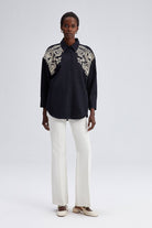 Model wearing black oversized shirt with cream embroidery on shoulders and front yoke.