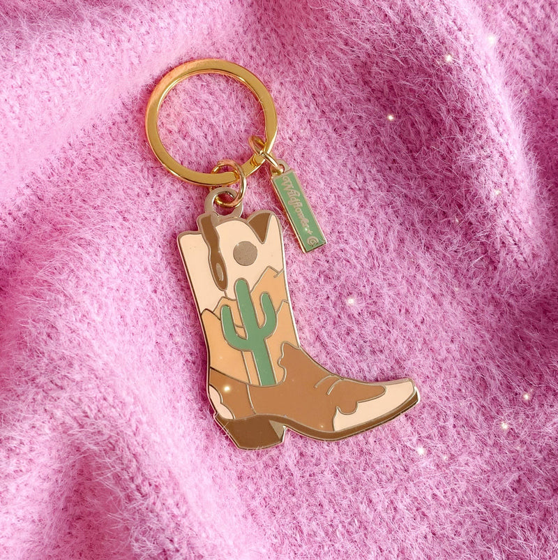 Desert Cowgirl Boot Enamel Keychain featuring a design with sunrise, clay-colored canyons, and a saguaro cactus.