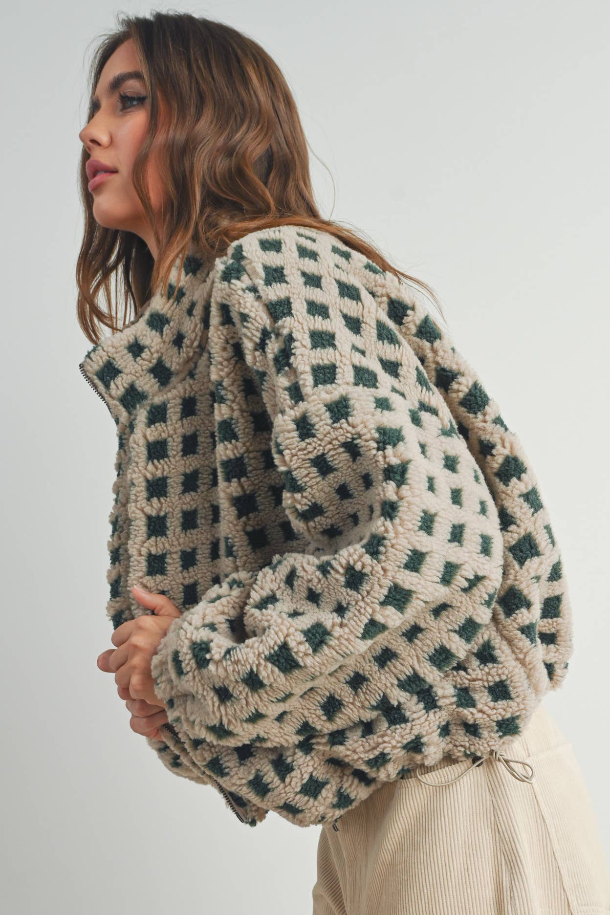 Woman wearing a beige and green checkered fleece jacket with a zip closure, paired with a white crop top and beige corduroy pants.