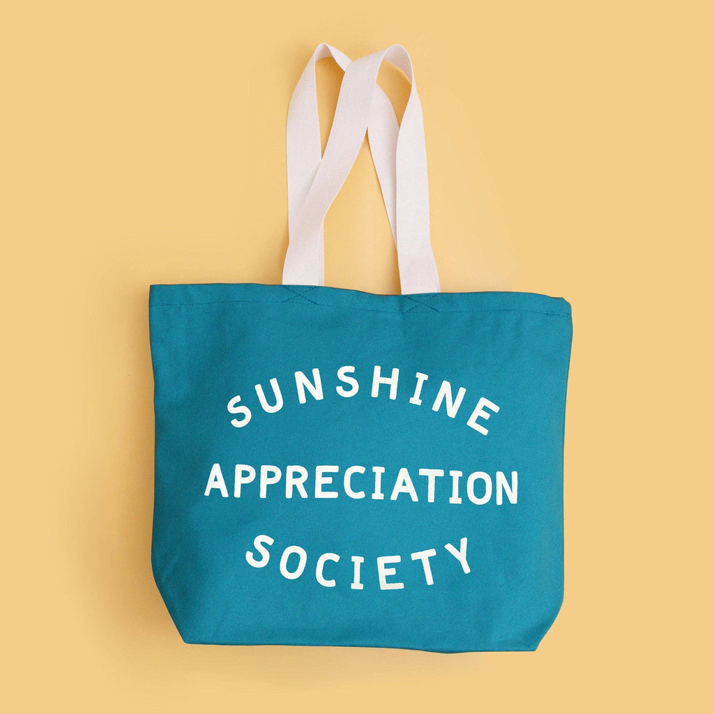 Teal tote bag with “Sunshine Appreciation Society” in white text, held by a person in a green dress at the beach