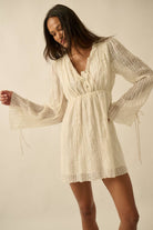 Woman wearing a cream mini dress with sheer long sleeves, button front, and tie details.